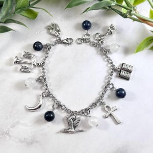 May include: A silver-toned charm bracelet featuring various metal charms including a scale of justice, an open book, an ankh, a crescent moon, a bird in flight, and an ink bottle, interspersed with dark blue and translucent white round beads.