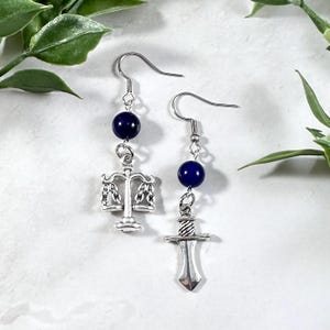 Puede incluir: Un par de pendientes de plata con cuentas de color azul oscuro. Un pendiente presenta un amuleto de balanza de la justicia, el otro un amuleto de espada.