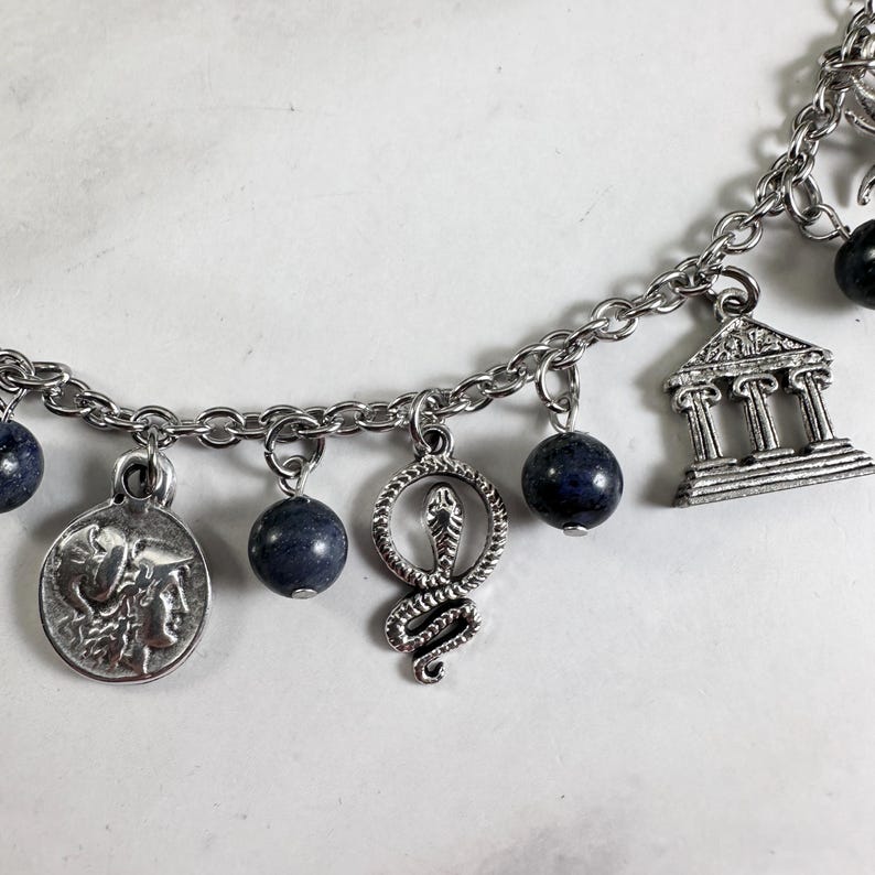 May include: A silver-toned charm bracelet featuring a series of dangling charms including a circular coin with a profile, a coiled snake, and a classical building with columns, interspersed with round, dark blue, speckled stone beads.