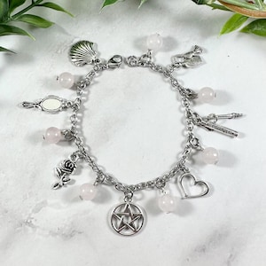 May include: A silver-toned charm bracelet featuring a variety of charms, including a seashell, mirror, rose, pentagram, heart, and other symbols. The bracelet also includes pink, round beads. The bracelet is displayed on a white surface with green foliage.