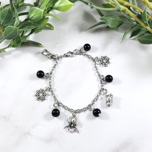 May include: A silver chain bracelet with black beads and charms. The charms include a spider, a web, and a spool of thread.