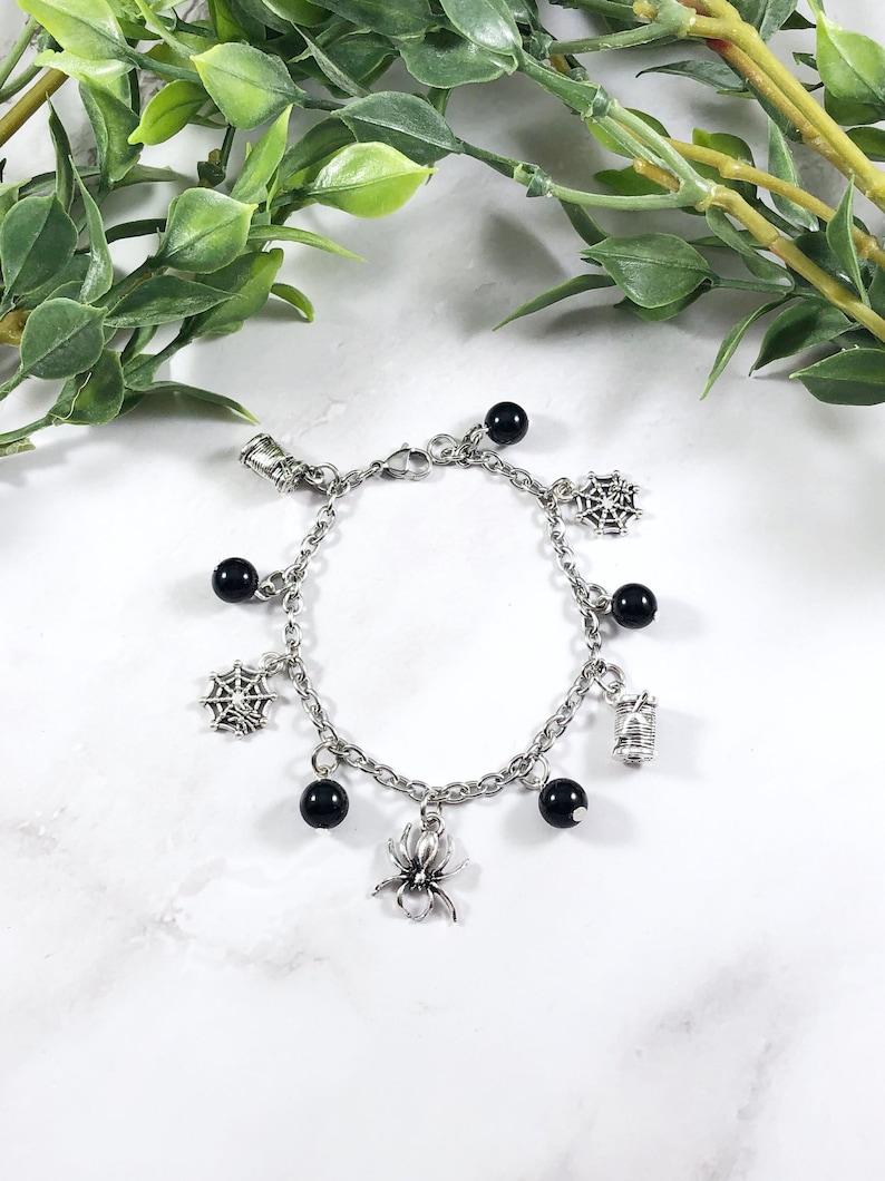 May include: A silver chain bracelet with black beads and charms. The charms include a spider, a web, and a spool of thread.