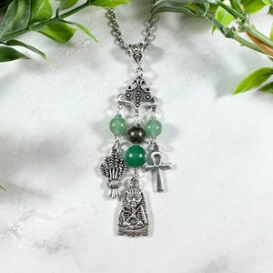 May include: Silver-toned pendant necklace featuring green gemstone beads and charms. The charms include a wheat sheaf, an ankh, and a pharaoh. The necklace has a decorative filigree design at the top and a chain.