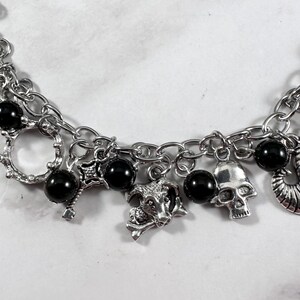 May include: Silver-toned charm bracelet featuring a variety of gothic-themed charms. The bracelet includes black beads, a skull, a goat head, and other decorative elements. The chain is made of linked metal.