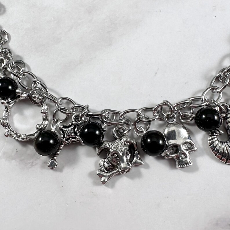 May include: Silver-toned charm bracelet featuring a variety of gothic-themed charms. The bracelet includes black beads, a skull, a goat head, and other decorative elements. The chain is made of linked metal.