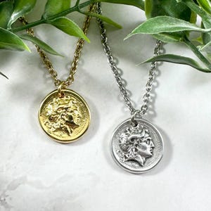 May include: Two coin pendant necklaces. One is gold-toned, the other silver-toned. Both feature a detailed relief of a classical figure on a round pendant. The necklaces hang from delicate chain links, with green foliage in the background.