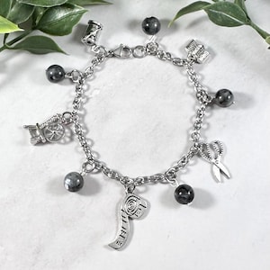 May include: A silver charm bracelet featuring a variety of sewing-themed charms, including a spool of thread, scissors, a thimble, and a measuring tape. The bracelet also includes several dark grey gemstone beads.
