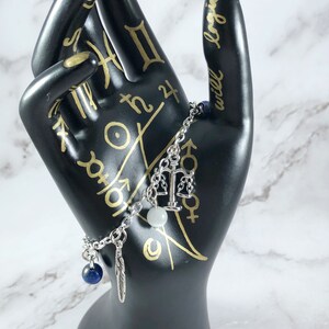 May include: A silver charm bracelet with a scale of justice charm, a white bead, a blue gemstone, and a feather charm.