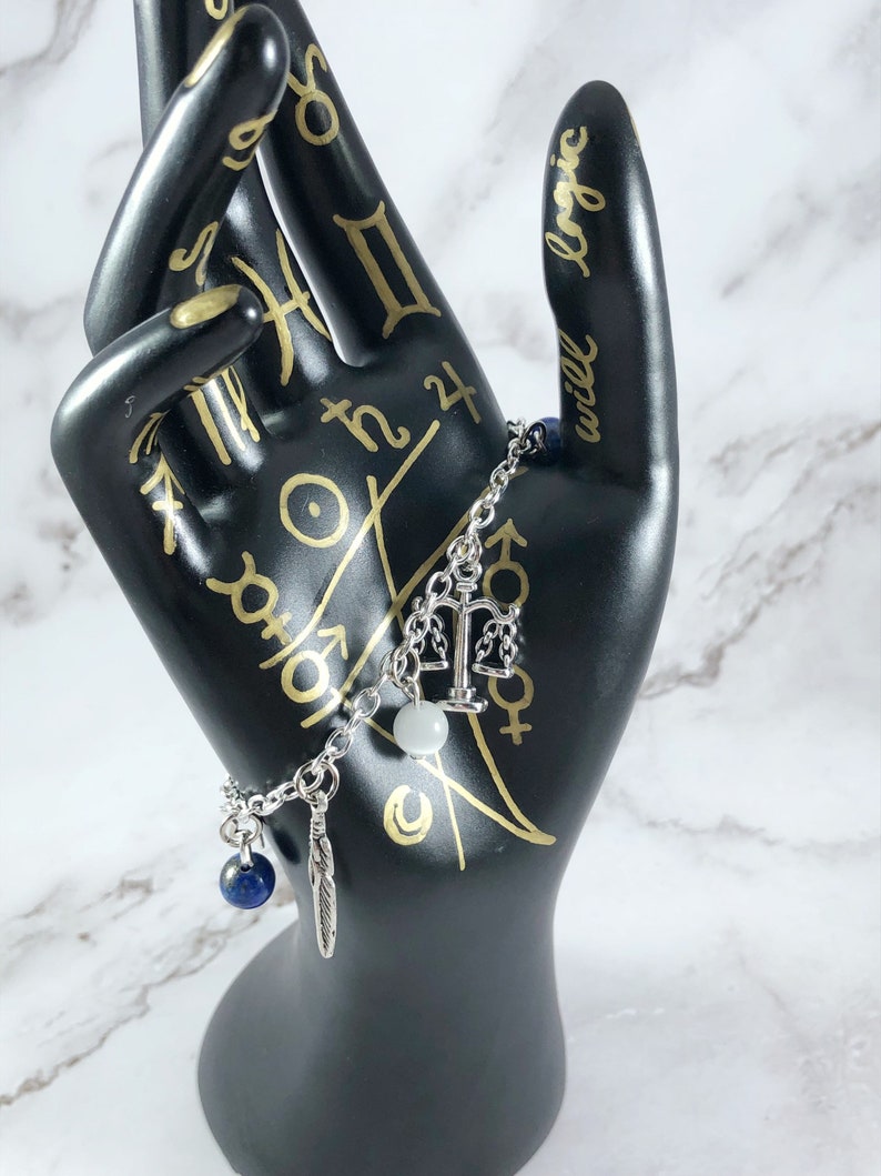 May include: A silver charm bracelet with a scale of justice charm, a white bead, a blue gemstone, and a feather charm.