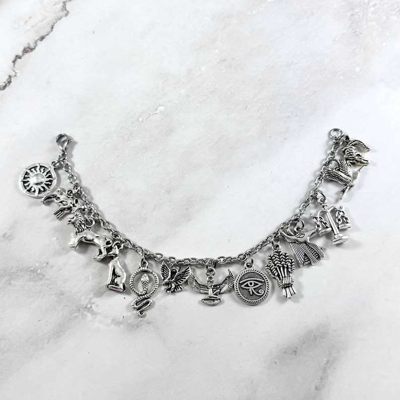 May include: Silver charm bracelet with various Egyptian symbols, including an ankh, a scarab beetle, a cobra, a sun disc, a winged sun, a lion, a goddess, a scale, a wheat sheaf, and the Eye of Horus.