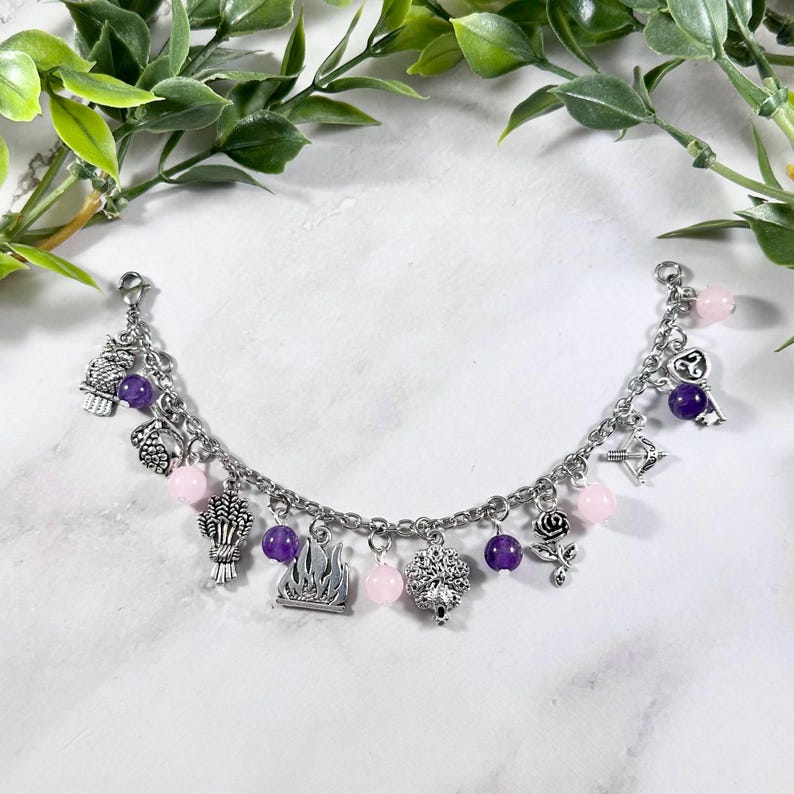 May include: A silver charm bracelet with pink and purple beads. The charms include a flame, a rose, a key, an arrow, a wheat bundle, and a tree.