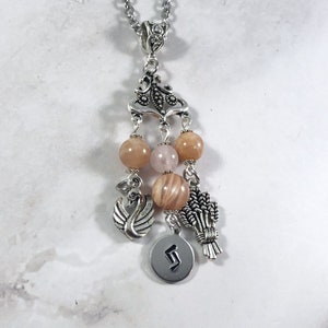 May include: A silver charm necklace with a delicate design, featuring a swan, wheat, and a round charm with the letter 'S' engraved on it. The necklace is adorned with pink and peach-colored beads.