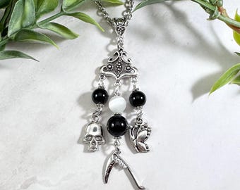 May include: A silver-toned pendant necklace featuring a decorative filigree bail, three dangling strands with black and white beads, a silver skull charm, a silver butterfly charm, and a silver scythe charm hanging from a metal chain.