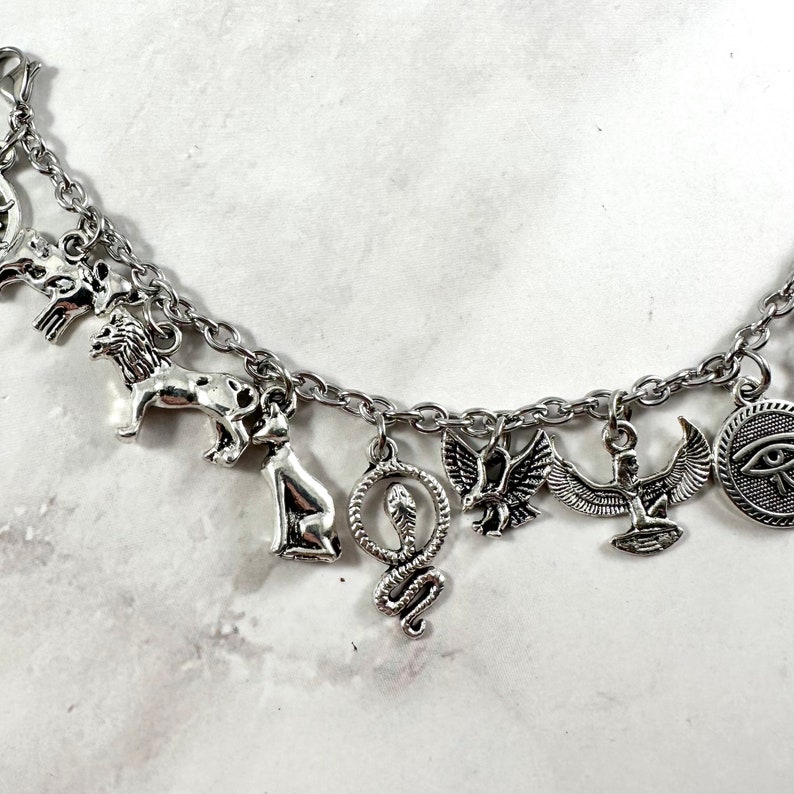 May include: Silver charm bracelet with multiple Egyptian-themed charms, including a lion, a snake, an ankh, a scarab beetle, and an eye of Horus.
