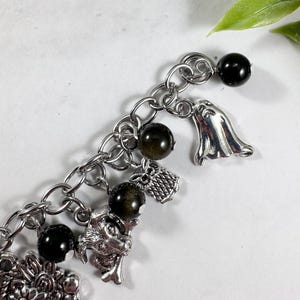 May include: A silver-toned charm bracelet featuring a variety of charms. The bracelet includes black spherical beads, a ghost charm, an owl charm, and a dragon charm. The bracelet is made of a chain link design.