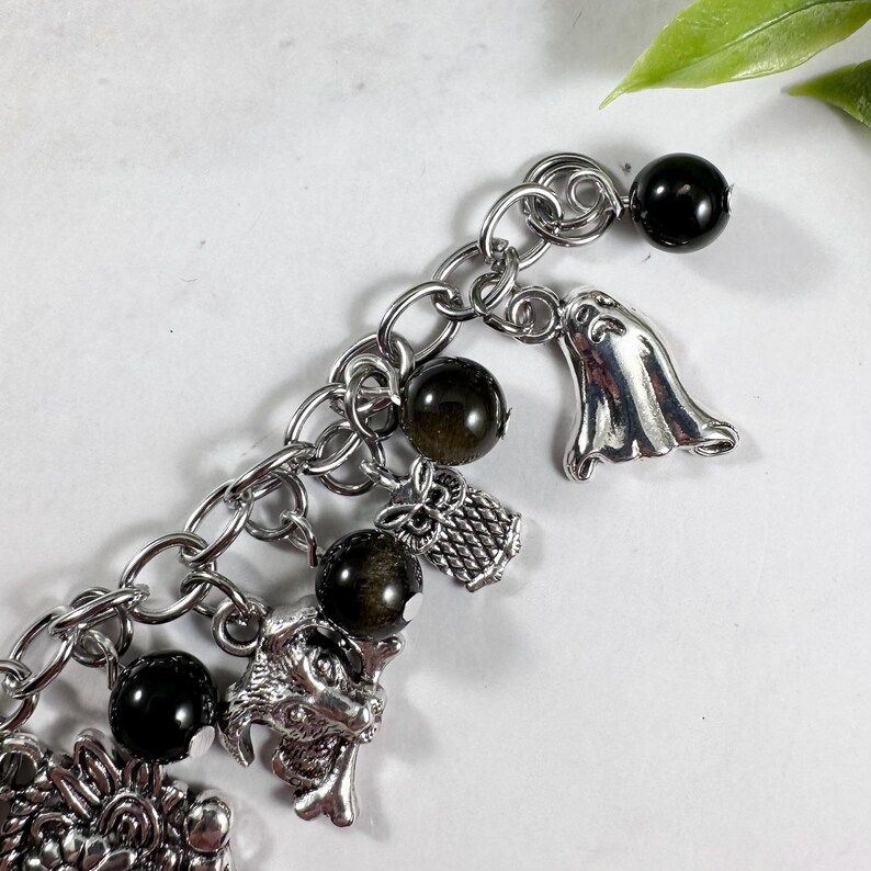 May include: A silver-toned charm bracelet featuring a variety of charms. The bracelet includes black spherical beads, a ghost charm, an owl charm, and a dragon charm. The bracelet is made of a chain link design.