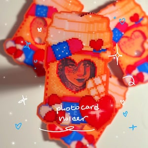 Pode incluir: Porta-cartões de fotos laranja com um design pixelizado. O suporte apresenta um recorte em forma de coração com um retrato no interior e é decorado com detalhes em vermelho, azul e branco. O texto "photo card holder" é visível.