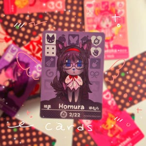 K&ouml;nnte beinhalten: Eine rechteckige Sammelkarte mit violettem Hintergrund und einer zentralen Illustration einer dunkelhaarigen Figur mit Katzenohren und Brille. Die Karte zeigt den Text Homura, 2/22 sowie verschiedene kleine Symbole wie einen Schmetterling.