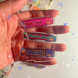 Op de afbeelding: Drie rechthoekige acryl bedels met anime-stijl illustraties en tekst. De bovenste bedel is roze met "Night Fever". De middelste bedel is rood en groen met "World Is Mine". De onderste bedel is blauw met "Cantarella".