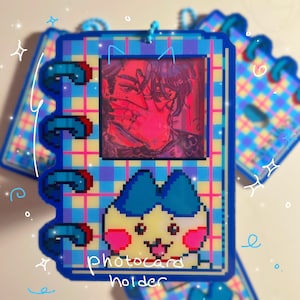 Pode incluir: Um porta-cartões fotográficos azul e amarelo com um design pixelizado. O porta-cartões apresenta um retrato e uma personagem de desenho animado. O texto "photocard holder" é visível na frente. O porta-cartões tem uma encadernação em espiral.