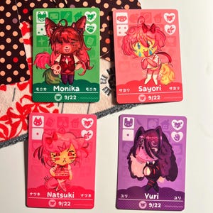 Puede incluir: Cuatro tarjetas coleccionables rectangulares con personajes de estilo anime llamados Monika, Sayori, Natsuki y Yuri. Cada tarjeta muestra un fondo de color, una ilustraci&oacute;n, iconos, el nombre en ingl&eacute;s y japon&eacute;s, y el texto 9/22.