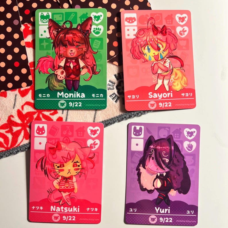 Puede incluir: Cuatro tarjetas coleccionables rectangulares con personajes de estilo anime llamados Monika, Sayori, Natsuki y Yuri. Cada tarjeta muestra un fondo de color, una ilustraci&oacute;n, iconos, el nombre en ingl&eacute;s y japon&eacute;s, y el texto 9/22.