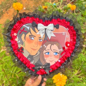 May include: Heart-shaped hand fan featuring an anime-style illustration of two people. The fan has a red and black ruffled trim, adorned with bows and embellishments. The handle is black.