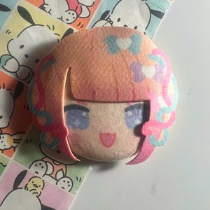 May include: A plush character head with peach-colored hair, blue and purple bows, and a smiling face with purple eyes. The character features pink hair accents and is positioned against a background of patterned paper featuring cartoon dog illustrations.
