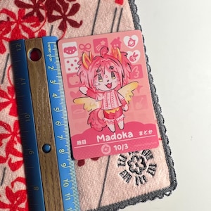 K&ouml;nnte beinhalten: Eine rechteckige Sammelkarte mit einer rosahaarigen anthropomorphen Figur namens Madoka mit gelben Fl&uuml;geln. Die Karte zeigt den Text Madoka, 10/3 und japanische Schriftzeichen. Ein Holzlineal zeigt eine H&ouml;he von etwa 9 cm an.