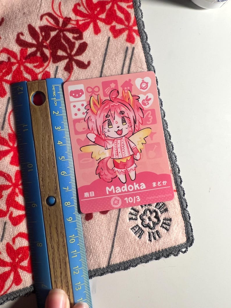 K&ouml;nnte beinhalten: Eine rechteckige Sammelkarte mit einer rosahaarigen anthropomorphen Figur namens Madoka mit gelben Fl&uuml;geln. Die Karte zeigt den Text Madoka, 10/3 und japanische Schriftzeichen. Ein Holzlineal zeigt eine H&ouml;he von etwa 9 cm an.