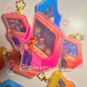 May include: Pink and clear acrylic keychain featuring a cartoon arcade game with a character playing. The game screen shows a group of cartoon characters and the text "Score Time 35". The keychain has the word "Shakers" written on the bottom.