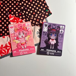 K&ouml;nnte beinhalten: Zwei rechteckige Sammelkarten mit Charakteren im Anime-Stil. Die linke Karte ist rosa mit einer Figur namens Madoka, die rechte Karte ist lila mit einer Figur namens Homura. Beide Karten zeigen verschiedene Symbole, Text und numerische Werte.