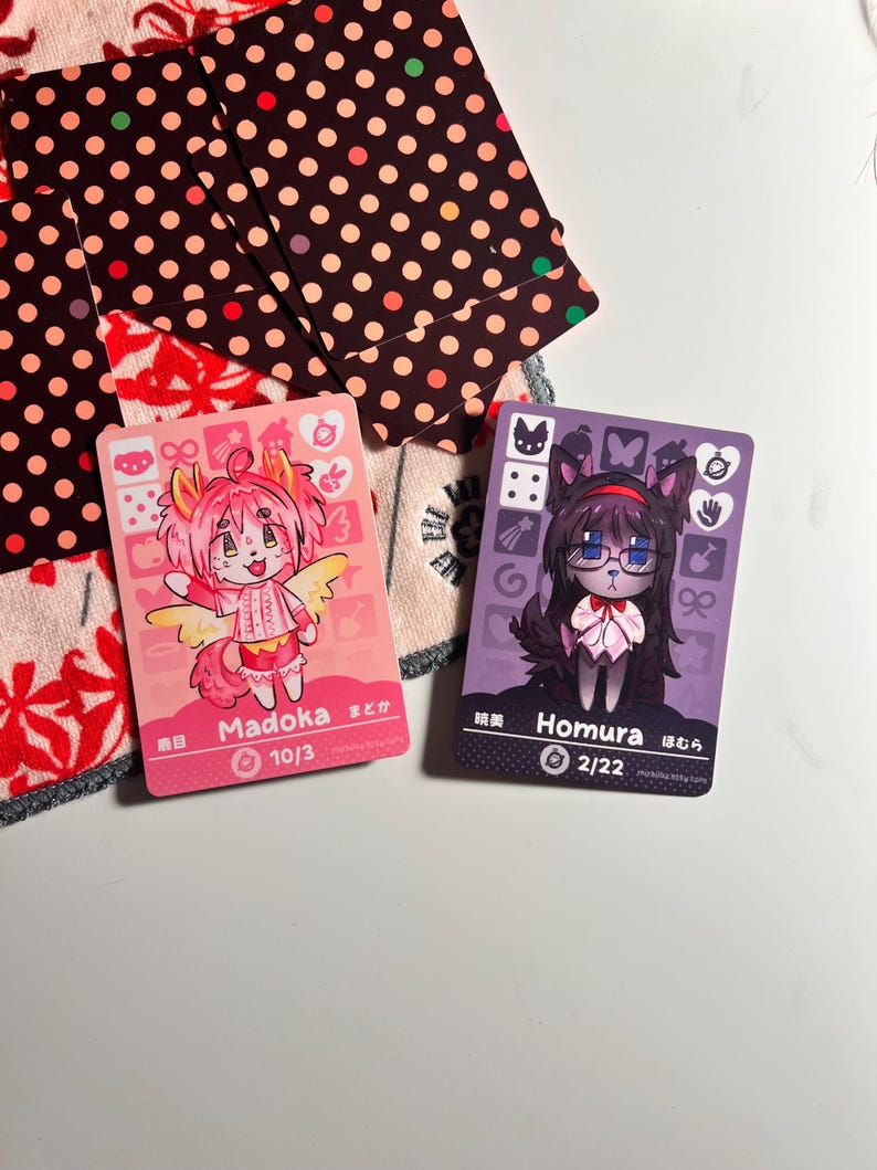 K&ouml;nnte beinhalten: Zwei rechteckige Sammelkarten mit Charakteren im Anime-Stil. Die linke Karte ist rosa mit einer Figur namens Madoka, die rechte Karte ist lila mit einer Figur namens Homura. Beide Karten zeigen verschiedene Symbole, Text und numerische Werte.