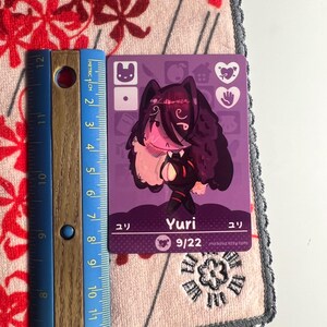 Puede incluir: Una tarjeta coleccionable rectangular de color morado con una ilustraci&oacute;n de personaje estilizado de cabello oscuro, etiquetada como Yuri con el texto 9/22. Una regla azul est&aacute; junto a la tarjeta, mostrando una altura de unos 9 cm.