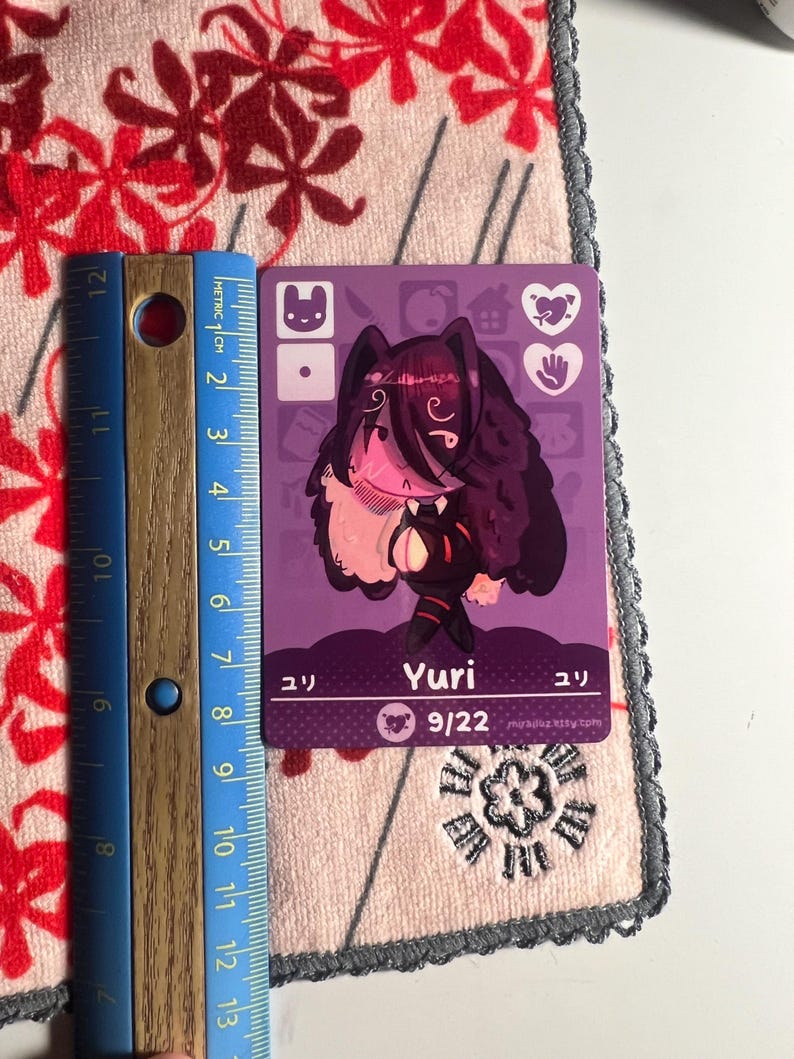 Puede incluir: Una tarjeta coleccionable rectangular de color morado con una ilustraci&oacute;n de personaje estilizado de cabello oscuro, etiquetada como Yuri con el texto 9/22. Una regla azul est&aacute; junto a la tarjeta, mostrando una altura de unos 9 cm.