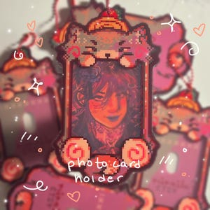 Pode incluir: Um porta-cartões fotográfico rosa com um design de gato pixelizado na parte superior e inferior. O suporte apresenta um retrato de uma pessoa com tons vermelhos e azuis. As palavras "photo card holder" estão impressas na frente.