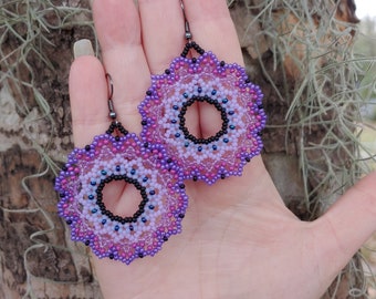 May include: A pair of purple and black beaded earrings with a mandala design. The earrings are round and have a hole in the centre.