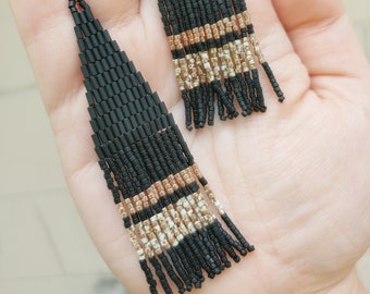 Pode incluir: Brincos longos de franjas de contas pretas e douradas. Os brincos têm forma triangular. A franja é feita de pequenas contas pretas com contas douradas e brancas intercaladas.