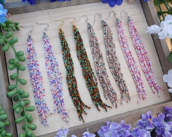 May include: Eight pairs of long, beaded earrings in various colours, including pink, purple, brown, and turquoise. The earrings are displayed on a wooden surface.