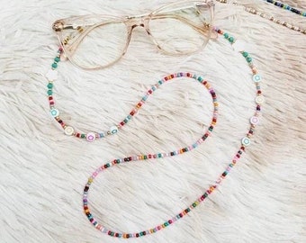 Puede incluir: Una cadena de gafas de cuentas de colores con pequeños abalorios blancos y de colores. La cadena está enroscada alrededor de unas gafas transparentes con montura marrón.