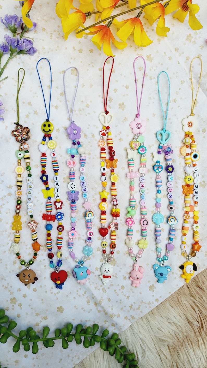 May include: Seven colourful beaded phone charms with different character charms at the bottom. The charms are in the shape of a bear, a heart, a blue bird, a white bunny, a koala, and a yellow puppy. The beads are in various colours, including red, yellow, blue, green, and pink. The charms are all strung on a thin string.