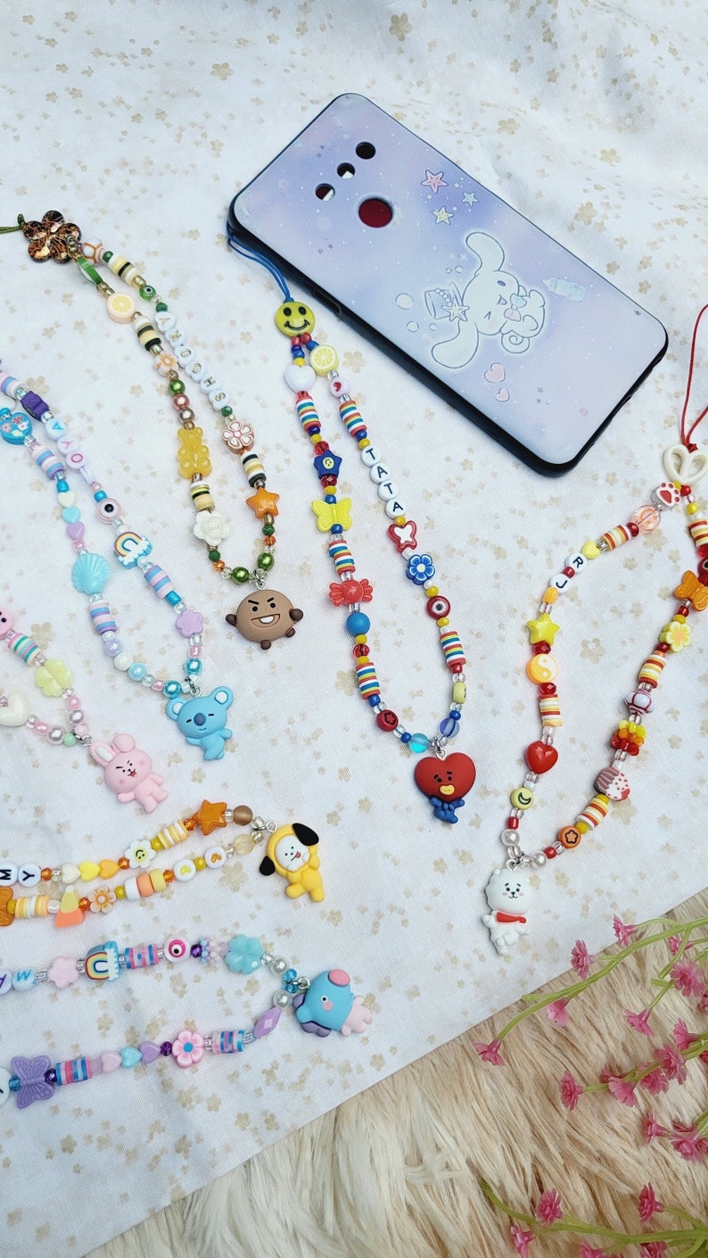 May include: A collection of colourful beaded phone charms featuring various cartoon characters, including a bunny, a koala, a bear, a dog, and a heart. The charms are attached to phone cases with a string of beads.
