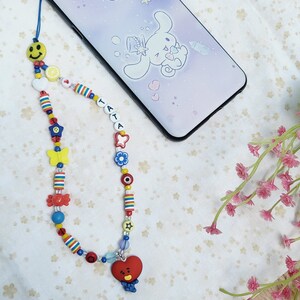 May include: A colourful beaded phone charm with a red heart-shaped charm featuring a cartoon face. The charm is attached to a phone case with a cartoon character design.