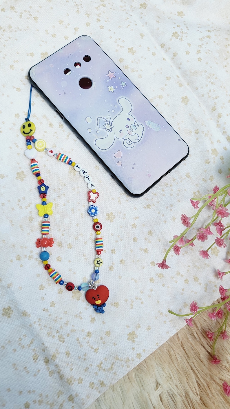 May include: A colourful beaded phone charm with a red heart-shaped charm featuring a cartoon face. The charm is attached to a phone case with a cartoon character design.