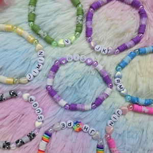 May include: A collection of colorful beaded bracelets. Each bracelet features a unique combination of colors and patterns, with some spelling out names or initials using white letter beads. The bracelets are displayed on a soft, multi-colored surface.