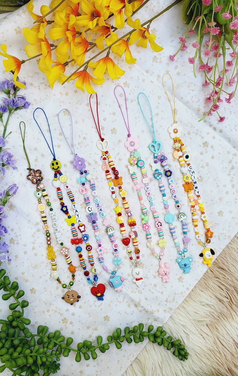 May include: Eight colourful beaded phone charms with different character charms at the bottom. The charms include a brown bear, a red heart, a blue bird, a white bunny, a grey koala, and a yellow dog. The beads are in various colours, including red, yellow, blue, green, and pink. The charms are spelled out with letter beads, including "JIMIN", "YOONGI", "NAMJOON", "TATA", "KOOKIE", "JHOPE", and "RM".