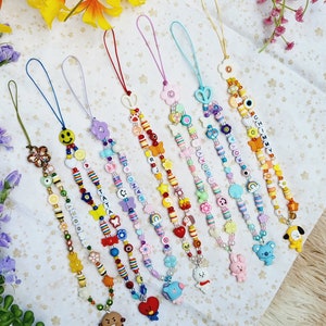 May include: A set of eight colourful beaded phone charms with different character charms. The charms include a flower, a smiley face, a butterfly, a rainbow, a heart, a bunny, a koala, and a dog. The beads are made of plastic and are strung on a string.