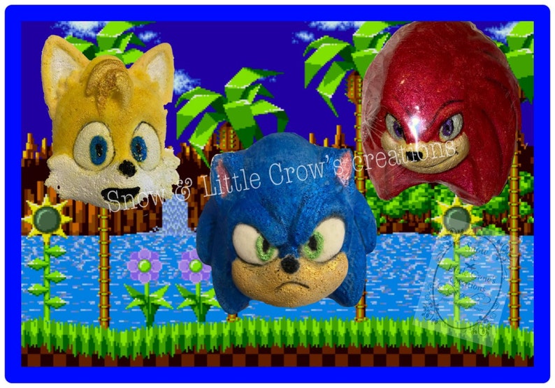 May include: Three bath bombs shaped like the heads of Sonic the Hedgehog, Tails, and Knuckles. The bath bombs are set against a pixelated background reminiscent of the Sonic the Hedgehog video game.