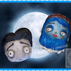 May include: Two handmade bath bombs shaped like the characters from the movie 'The Nightmare Before Christmas'. The bath bomb on the left is gray and white and shaped like Jack Skellington. The bath bomb on the right is blue and white and shaped like Sally. The bath bombs are in front of a large, full moon.