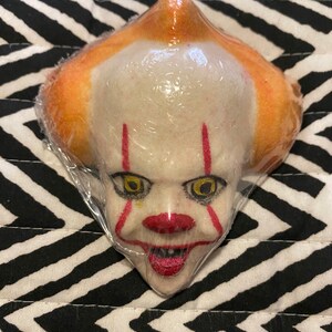 May include: A bath bomb shaped like Pennywise the clown from the movie "It". The bath bomb is white with red stripes and has a yellow and black eye. The bath bomb is wrapped in plastic.